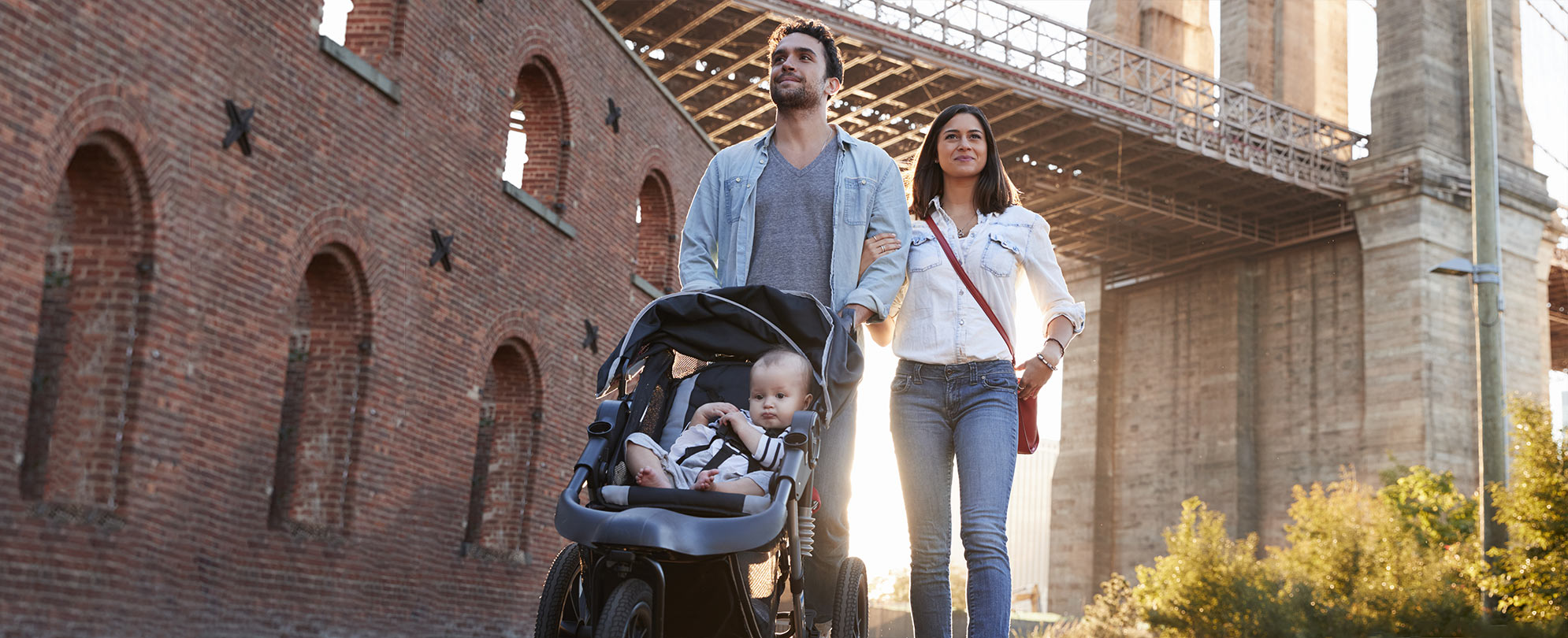 A young couple walking under a bridge while pushing a baby stroller with the bright sun behind them.