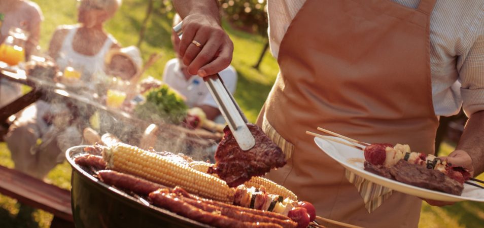 A man holds a plate of steak and vegetables in one hand while flipping a steak with a pair of tongs above a hot grill.
