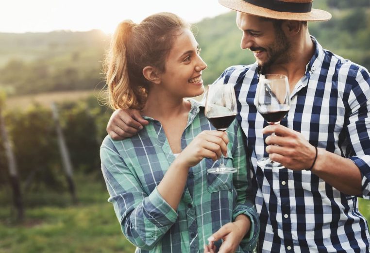 A couple in their 30s clink wine glasses full of red wine in a sunny vineyard