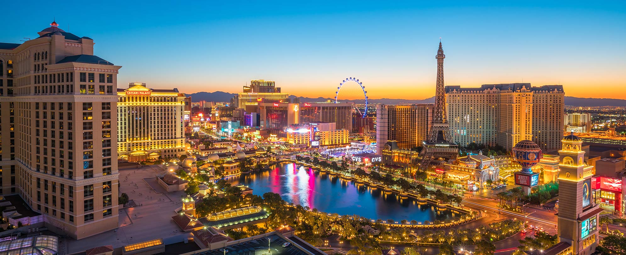 Aerial photo of Las Vegas Strip at dusk featuring the Paris Hotel replica Eiffel Tower, lake in front of Bellagio Hotel, Caesar's Palace, and view of High Roller observation wheel.