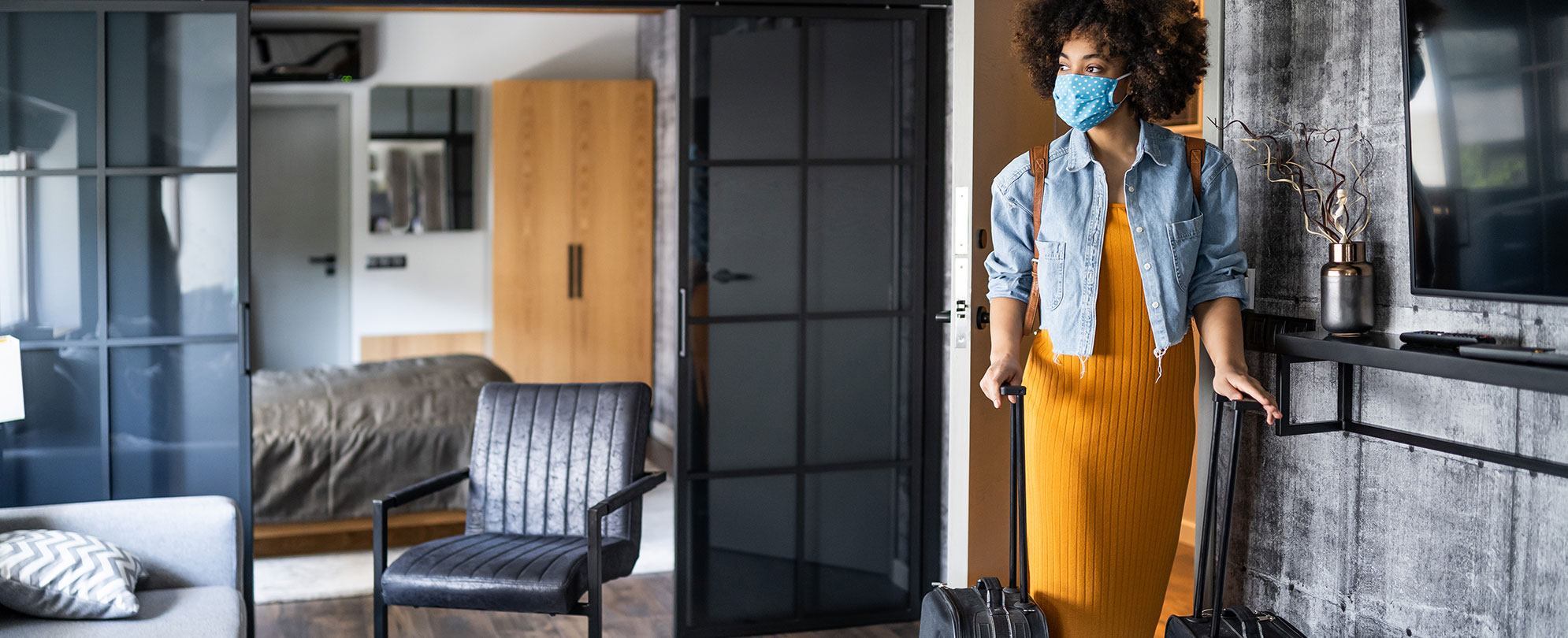 A woman wearing a face mask and yellow dress is rolling her suitcase into her resort suite. 