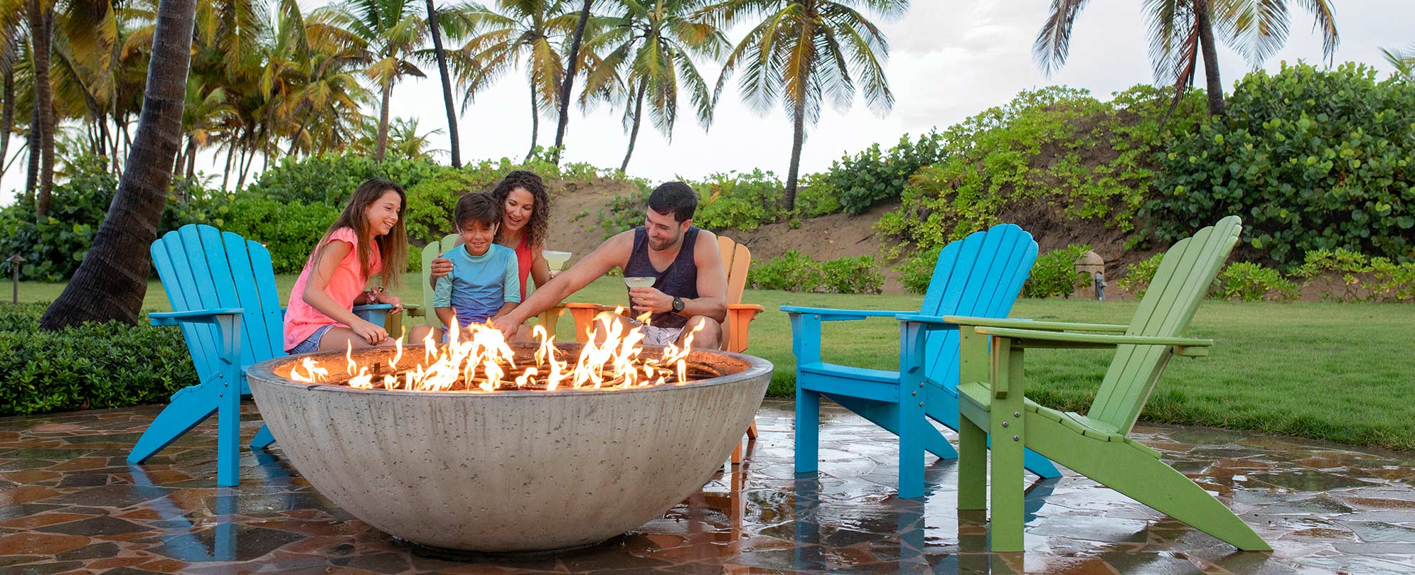 A family of four sitting in colorful wooden beach chairs around a large firepit at a Margaritaville Vacation Club resort. 