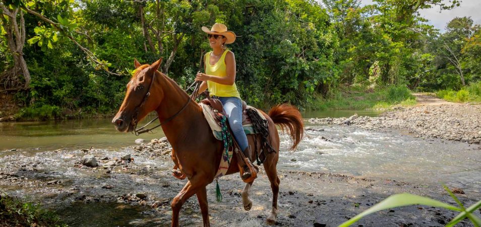 A woman wearing a yellow tank top and a wide-brimmed hat riding a horse through a rocky river. 