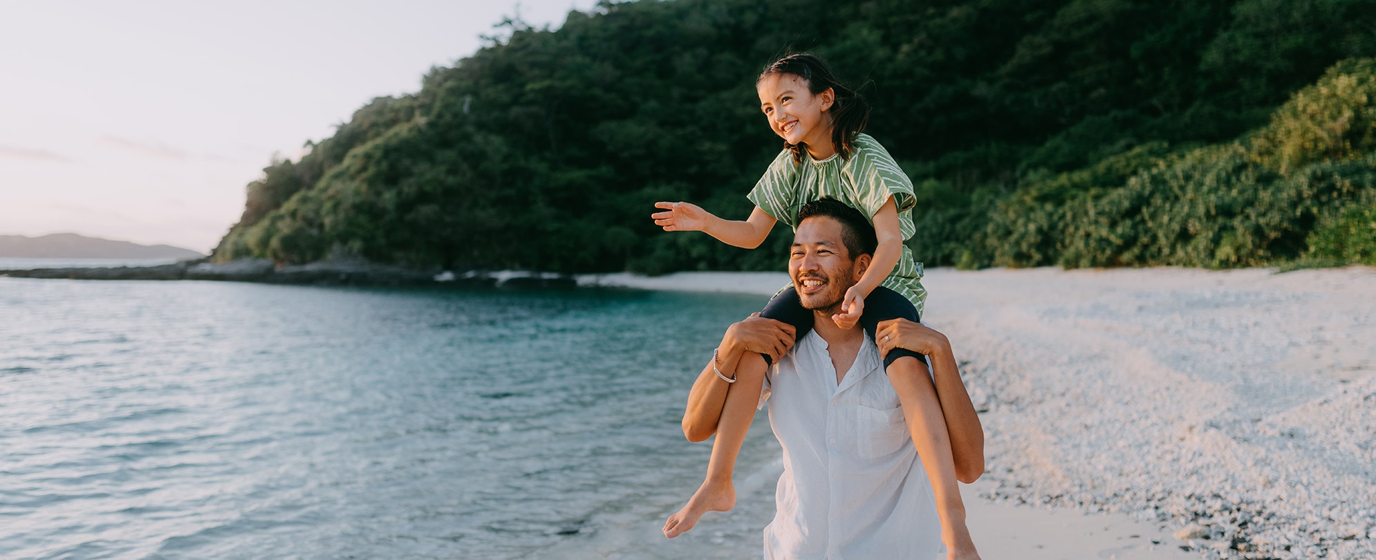 Father carries his daughter on her shoulders along the beach with the ocean and trees in the background.