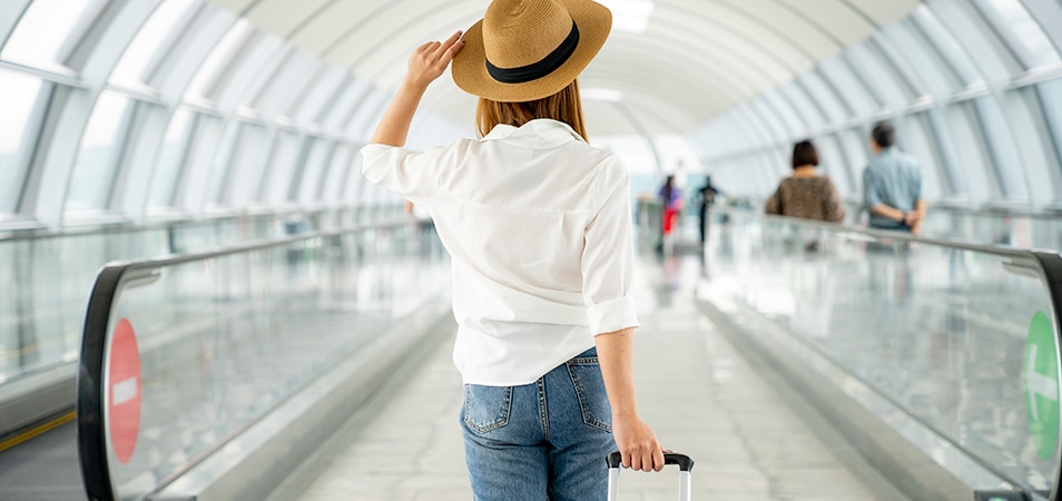 Woman holding her hat on her head and facing forward on a moving walkway at an airport.
