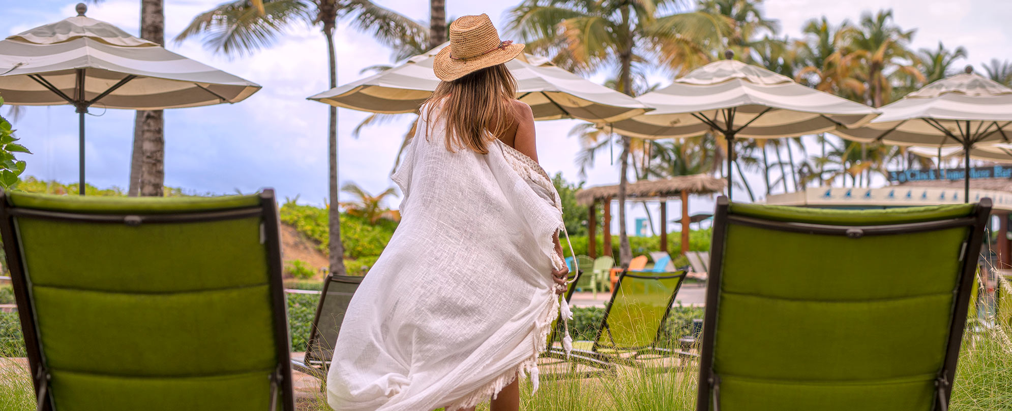 A woman with blonde hair wearing a hat and swimsuit cover-up walking to the Margarita Vacation Club resort pool. 