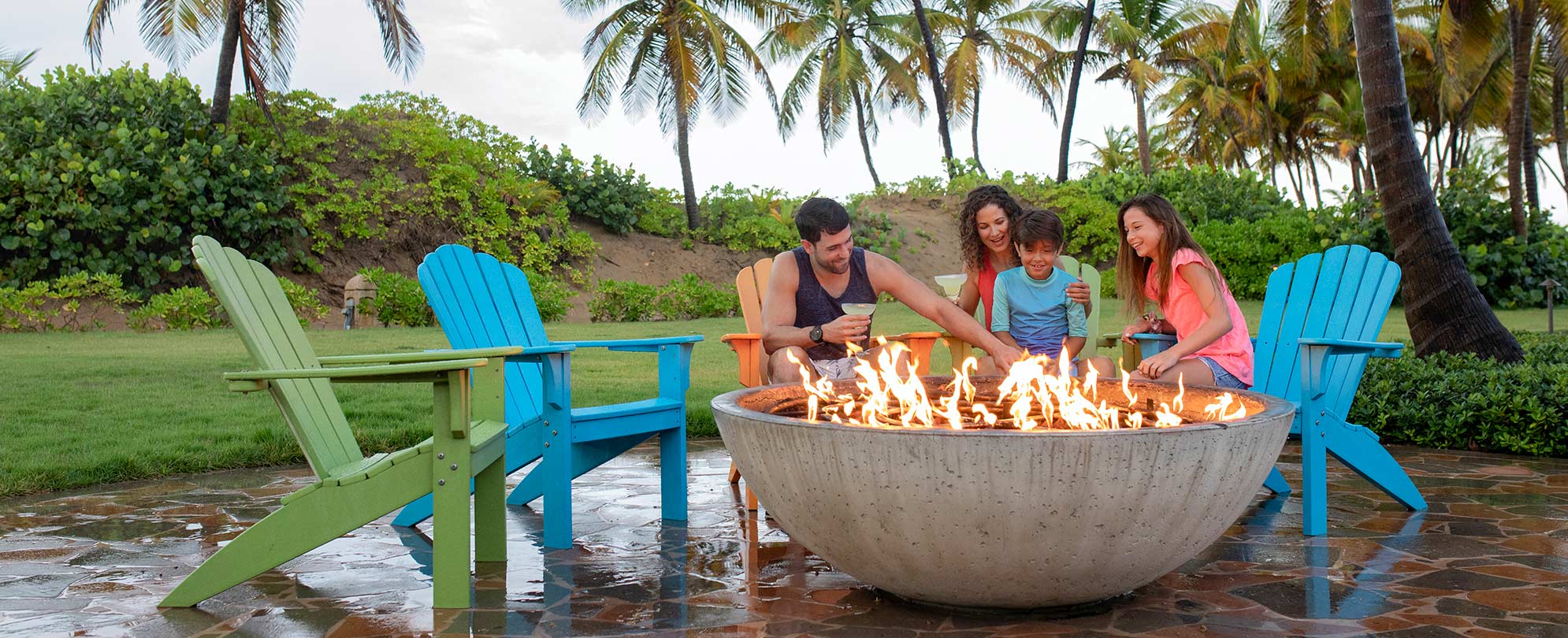 A family sits in adirondack chairs around a large round, stone firepit at a Margaritaville Vacation Club resort.