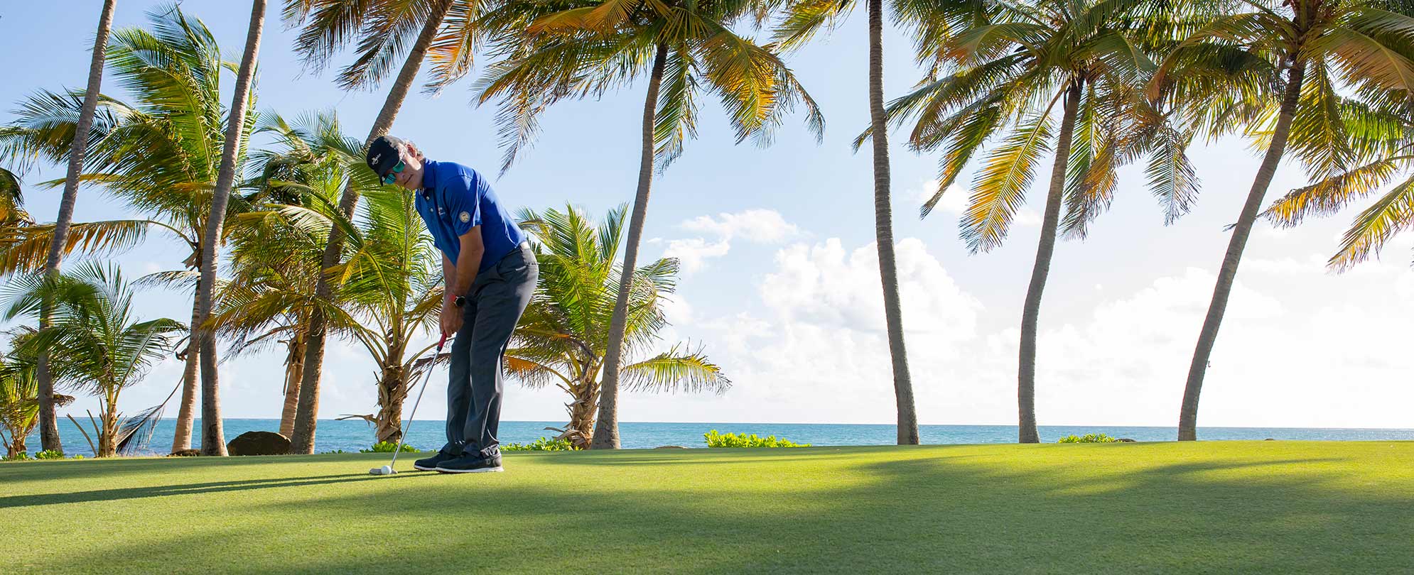 Man in a blue polo putting a golf ball at an oceanfront golf course near a Margaritaville Vacation Club resort.