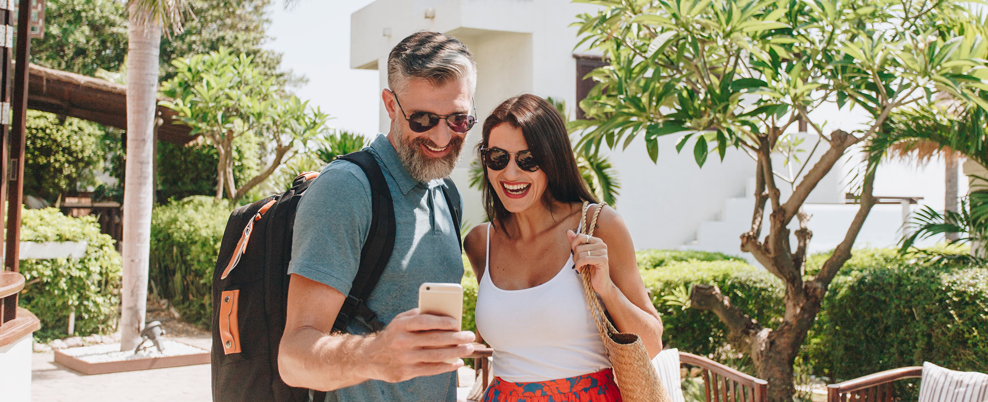 A man and a woman, both smiling and wearing sunglasses, stand in a sunny courtyard looking at something on the man's cell phone.