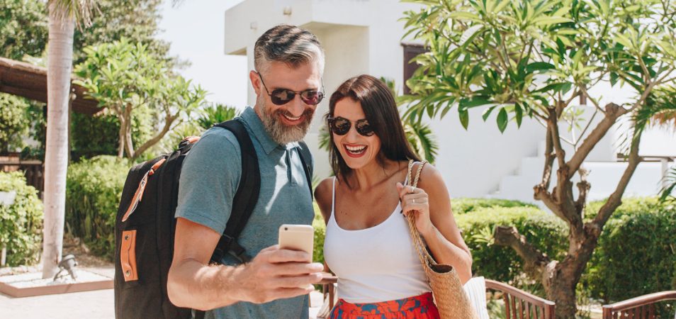 A man and a woman, both smiling and wearing sunglasses, stand in a sunny courtyard looking at something on the man's cell phone.