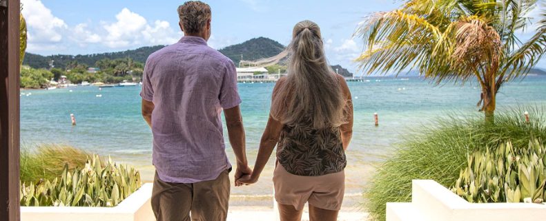 A couple holding hands looks out at the ocean from an outdoor pathway at a Margaritaville Vacation Club resort.
