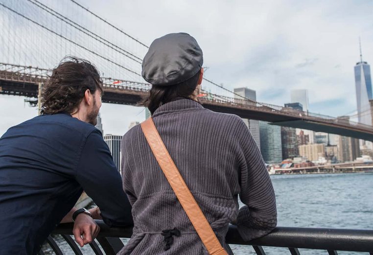 Man and woman leaned over railing, taking in the view of the New York City skyline and Brooklyn Bridge.