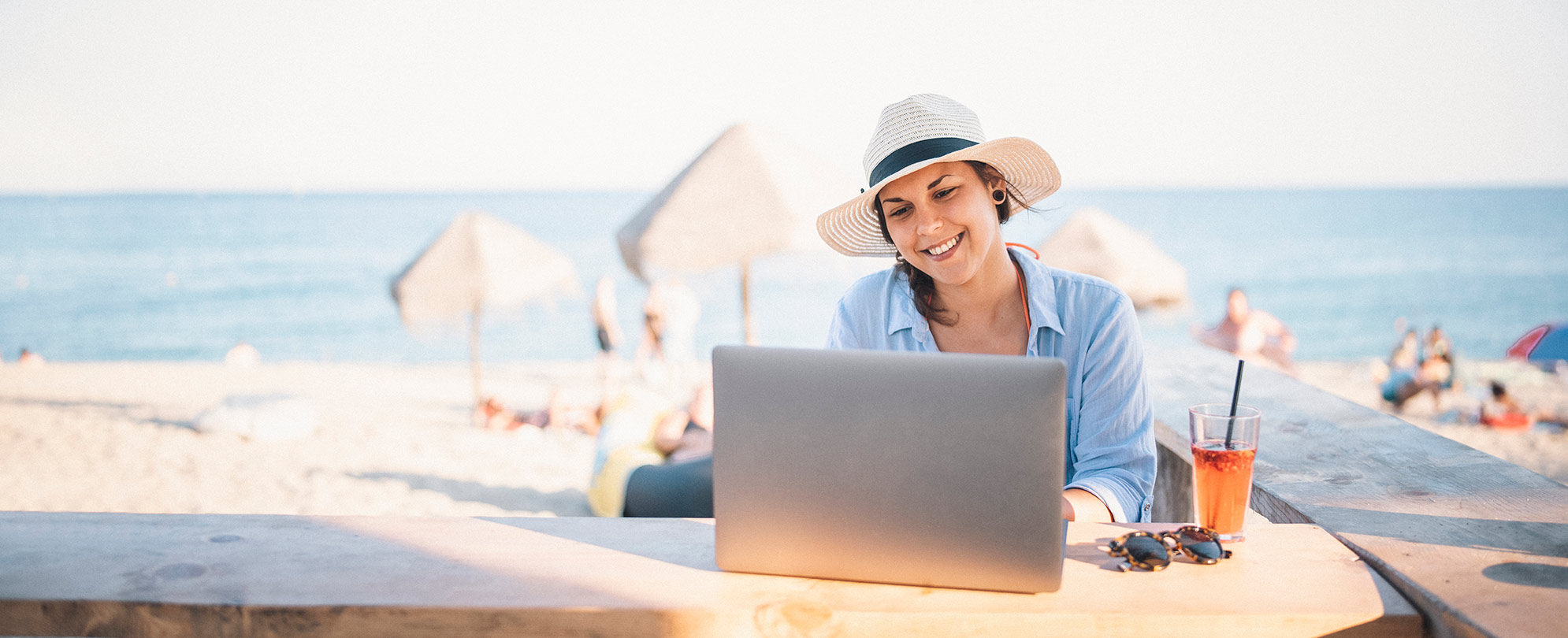 A smiling woman working remotely from a outdoor bar with the beach in the background.