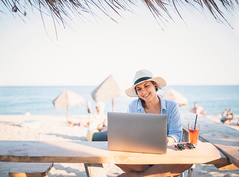 A smiling woman working remotely from a outdoor bar with the beach in the background.