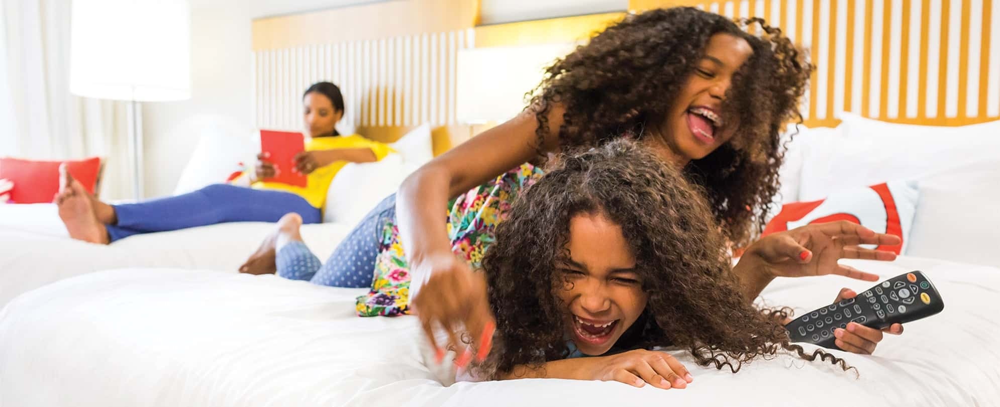 Two smiling young girls with dark, curly hair wrestle for remote on the bed of a Margaritaville Vacation Club Resort suite.