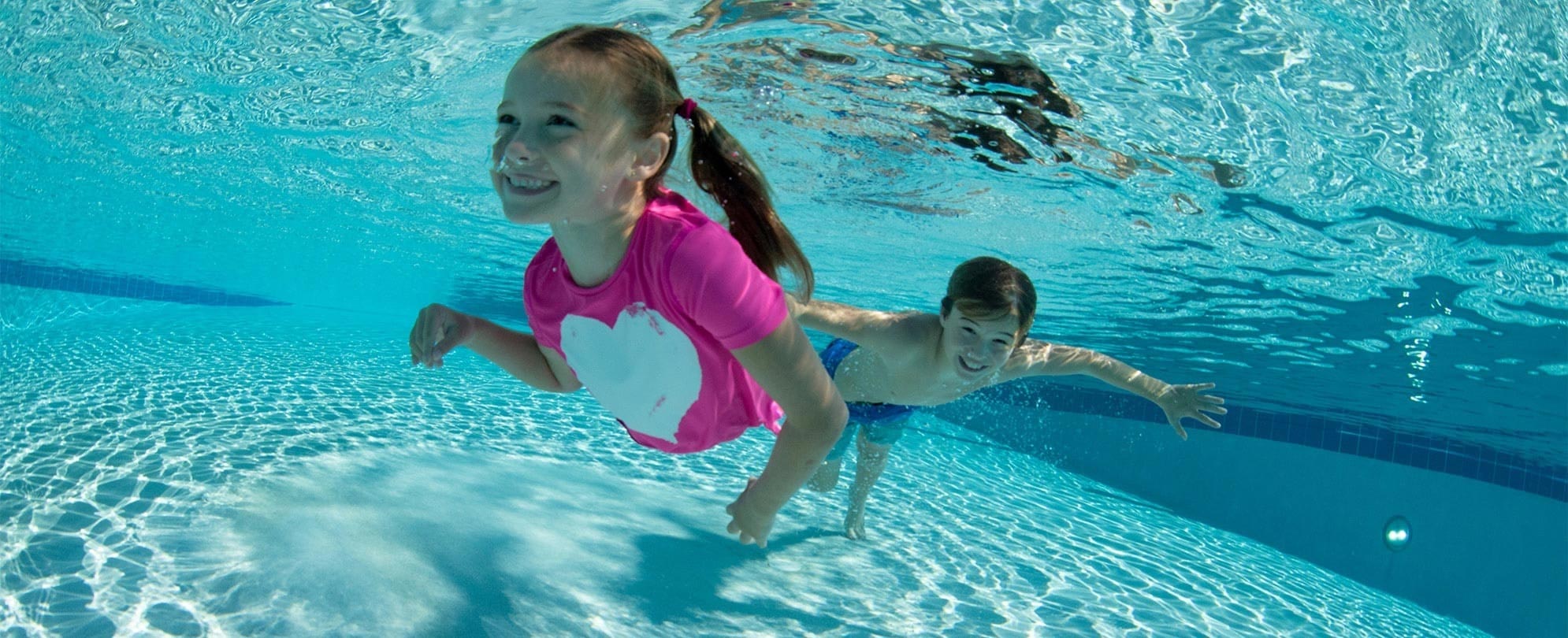 A smiling young girl and boy swim underwater in a pool while enjoying vacation.