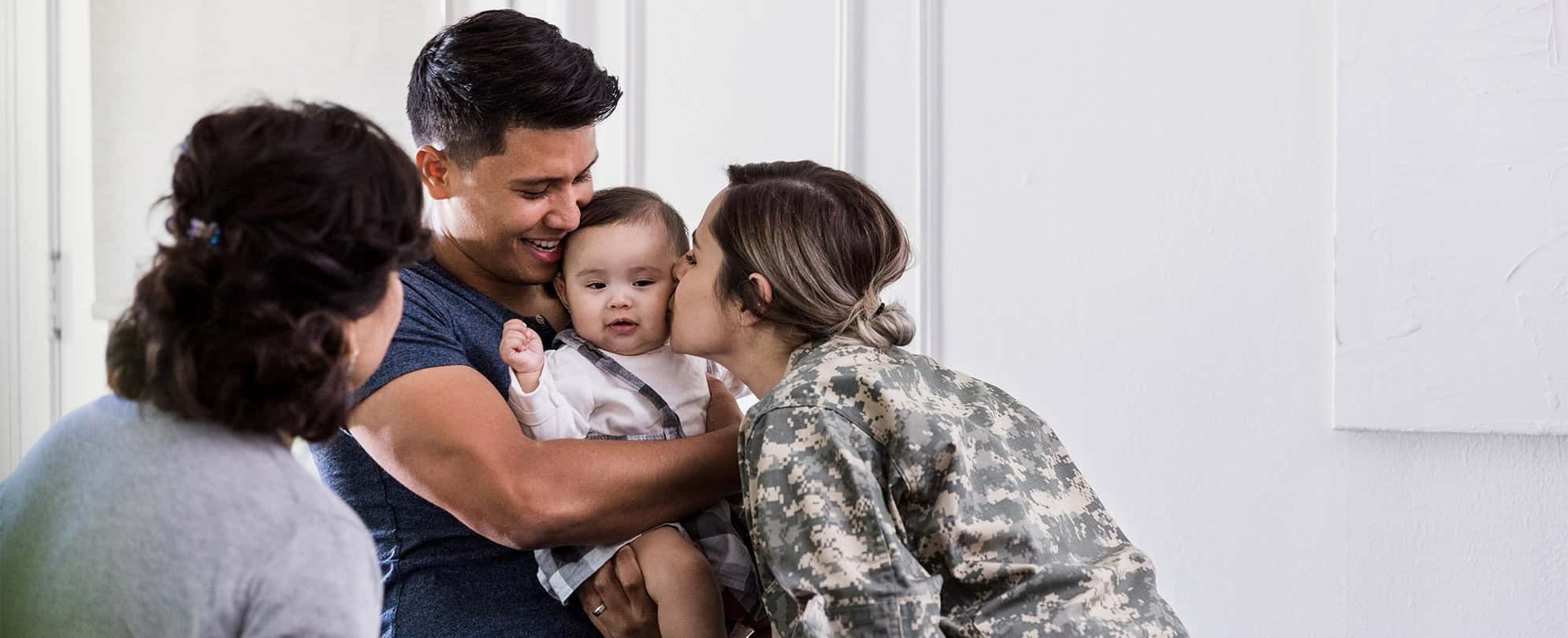 A grandmother lovingly watches a father holding baby while a military mom leans over to kiss the child on the cheek.