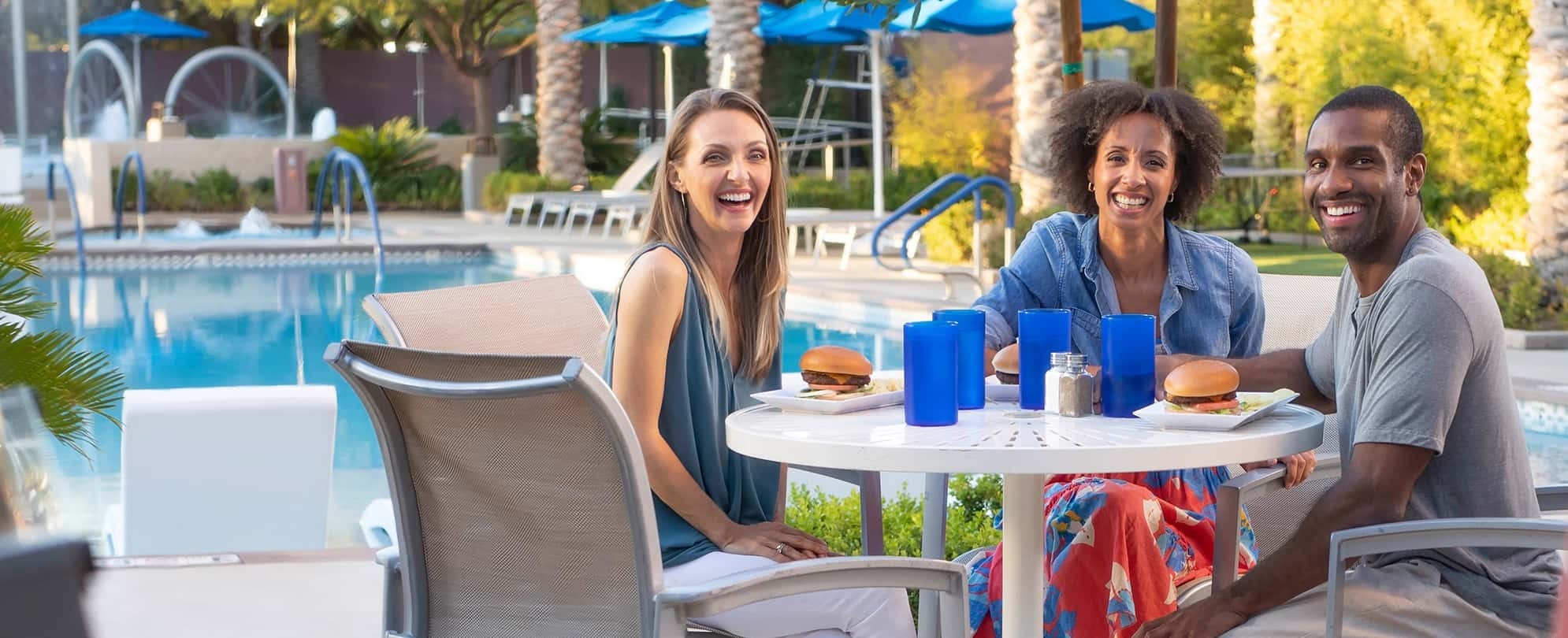 Three smiling adults enjoy burgers and drinks in blue cups at a poolside table of a Margaritaville Vacation Club resort.