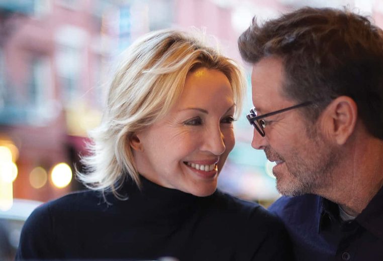 A man wearing glasses and woman in a black turtleneck shirt smile lovingly at each other.