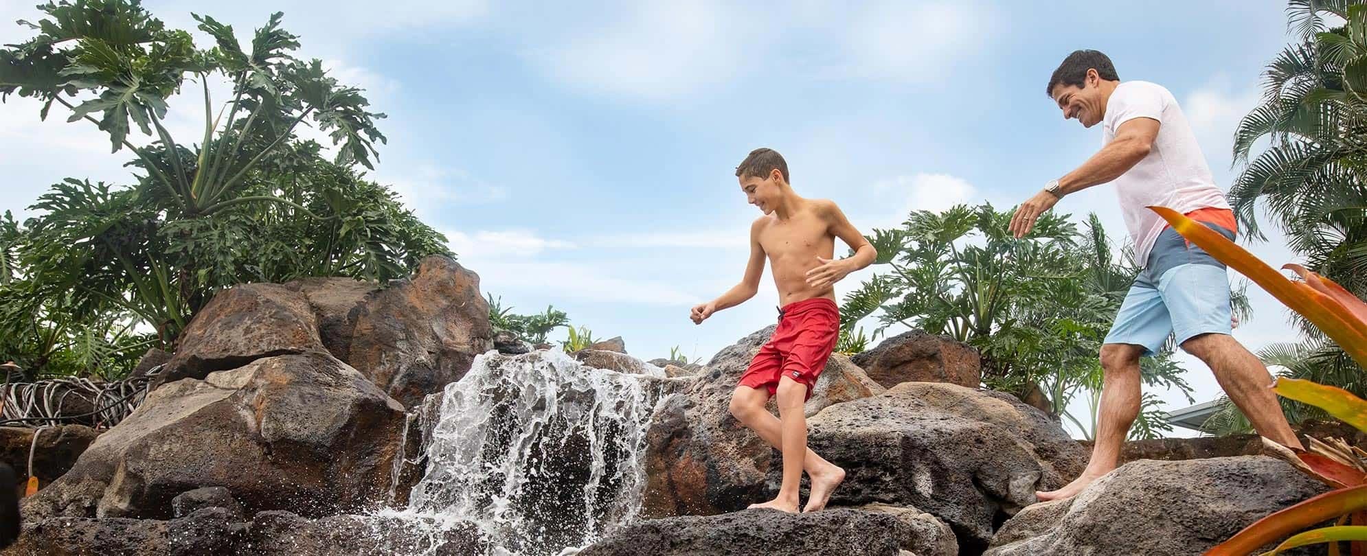 Dad and preteen boy walk on rocks in front of a waterfall during a tropical vacation.