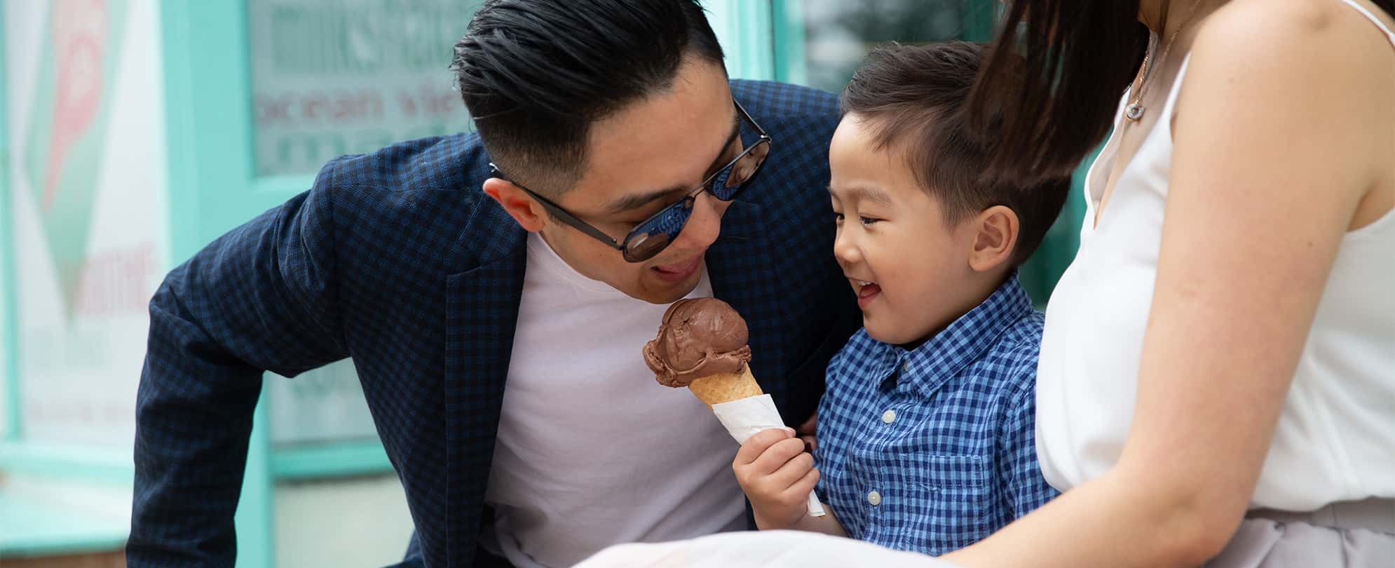 Young boy holding chocolate ice cream cone sits between mom and dad as the dad leans in to take a big bite of ice cream.