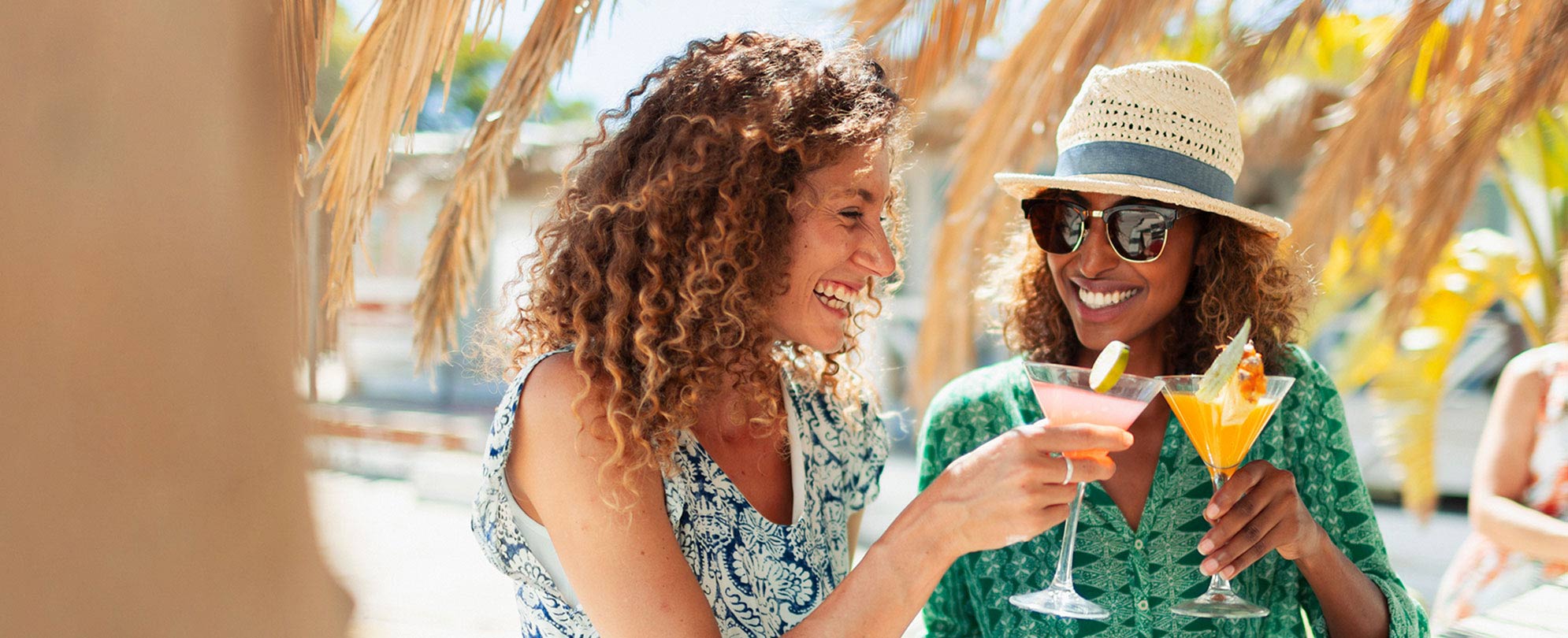 2 smiling women sitting under palm trees and clinking cocktail glasses together in a toast 