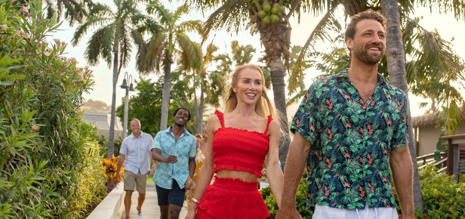 Men and women wearing bright, summer clothes, walk along a path surrounded by palms at a Margaritaville Vacation Club resort.
