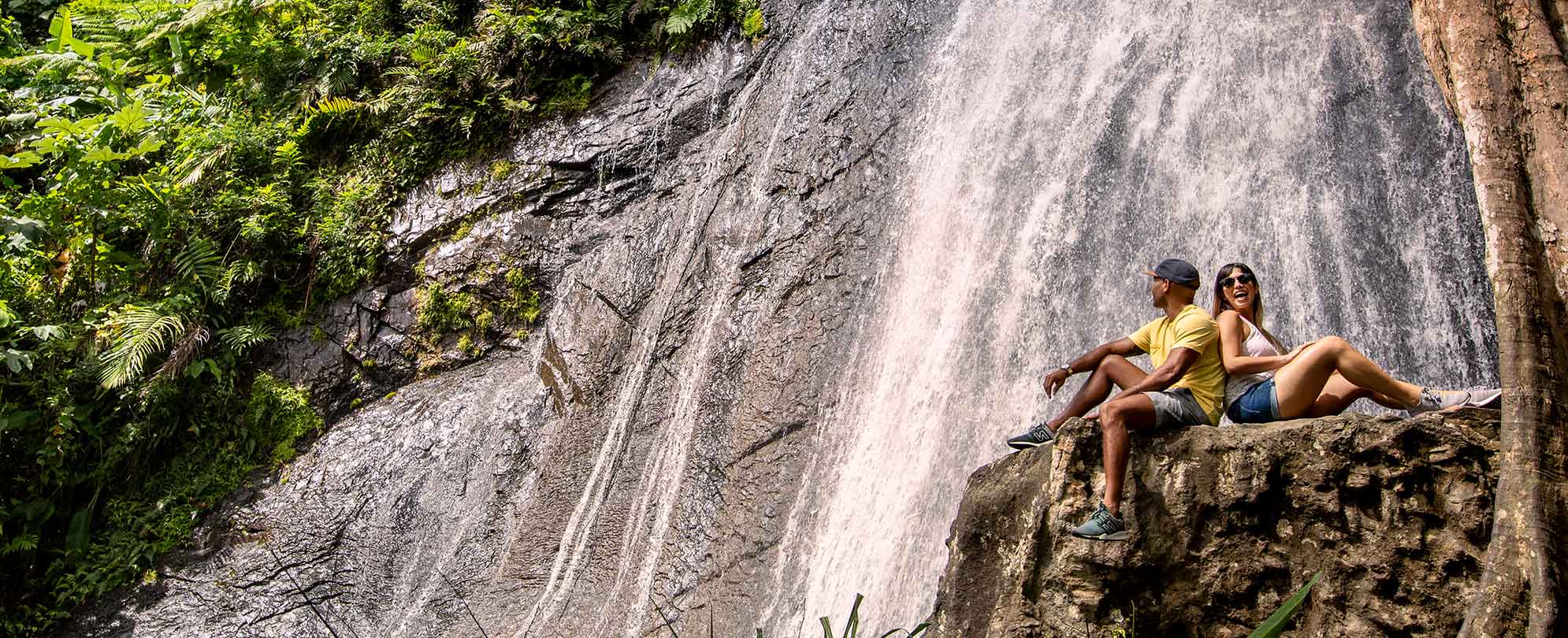 Man and woman sit back-to-back on a rock in front of a waterfall as they explore top things to do in Rio Mar, Puerto Rico.