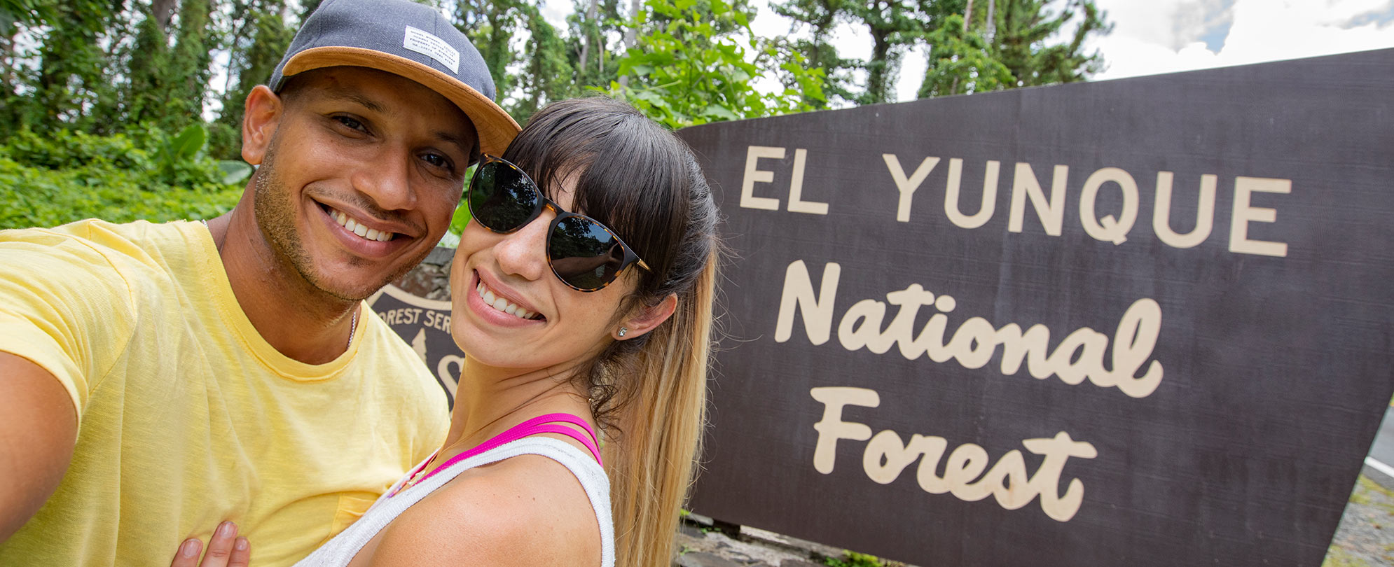 Man in hat and woman in sunglasses pose for a selfie in front of the El Yunque National Forest in Rio Grande, Puerto Rico.