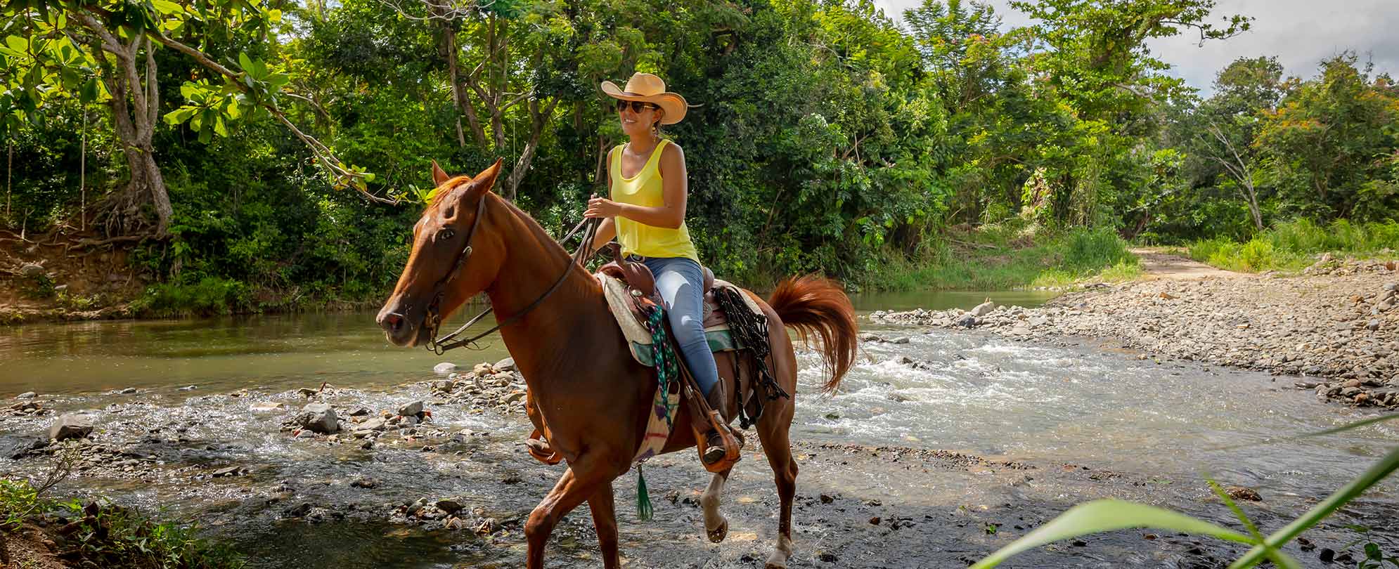 A woman rides a horse through a stream, enjoying outdoor adventures in Puerto Rico during her Margaritaville vacation.