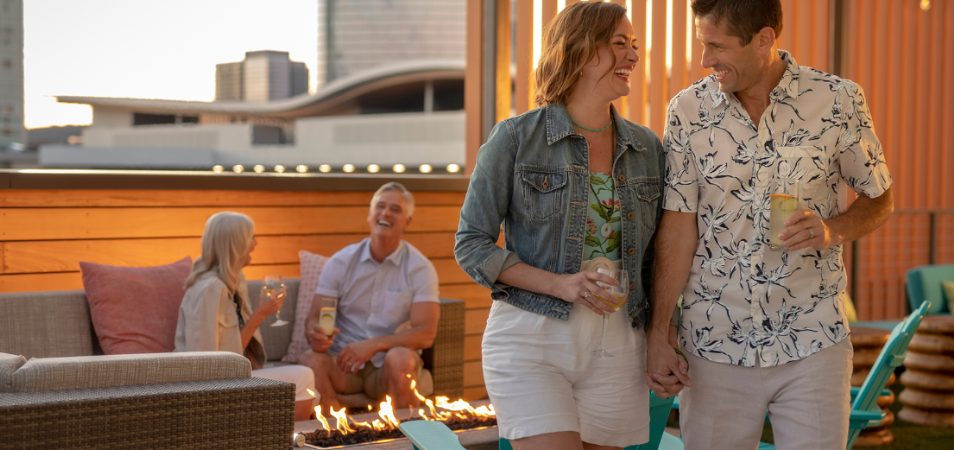 A couple standing and laughing with drinks in hand with a couple in the background sitting and laughing near the timeshare resort fire pit. 