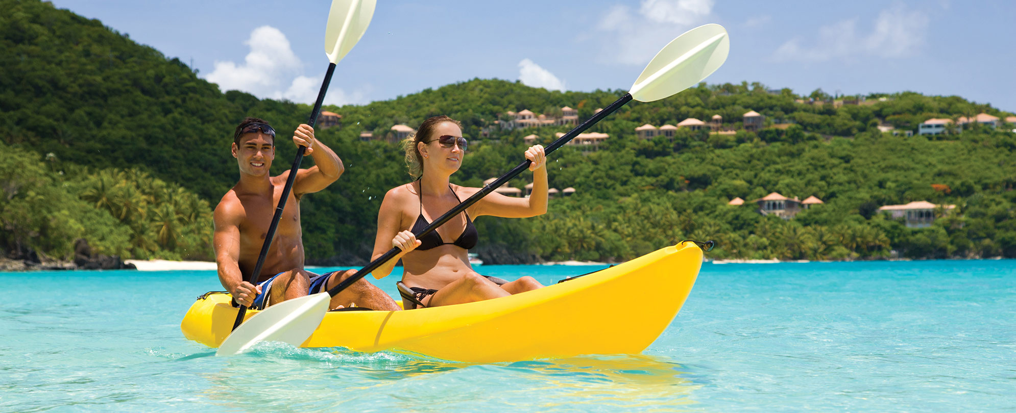 A man and woman paddle a double kayak in a tropical vacation destination.