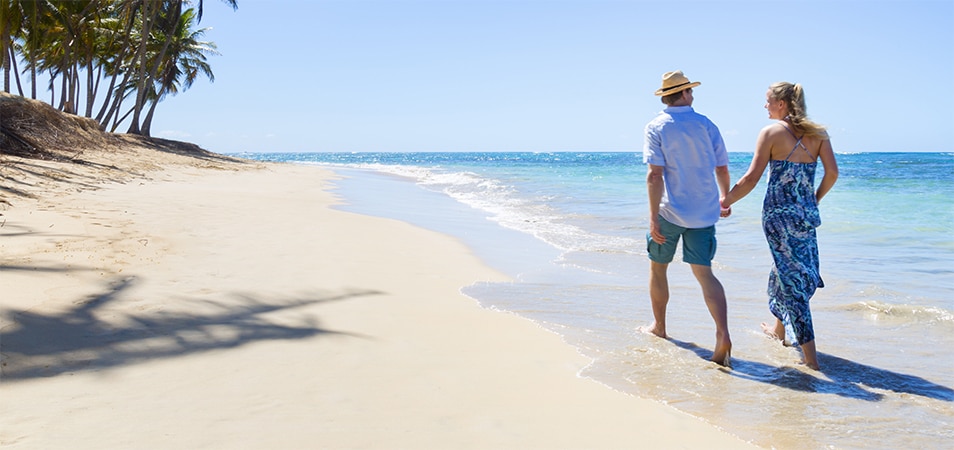 A man and woman holding hands and walking down the beach on their Margaritaville vacation.