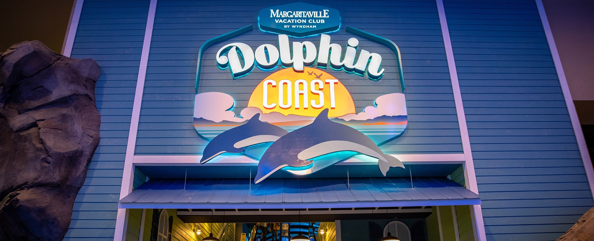 View of a large Dolphin Coast sign with a sun setting into ocean waves and two animated dolphins below the Margaritaville Vacation Club by Wyndham logo in the new gallery of the Georgia Aquarium.