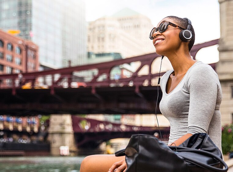 A woman sitting by a river flowing through a city listens to music on her headphones