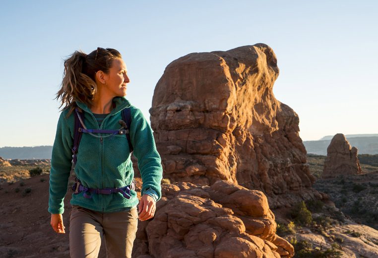 A woman in hiking gear looks back over her shoulder at large red rock formations while exploring the American Southwest.	