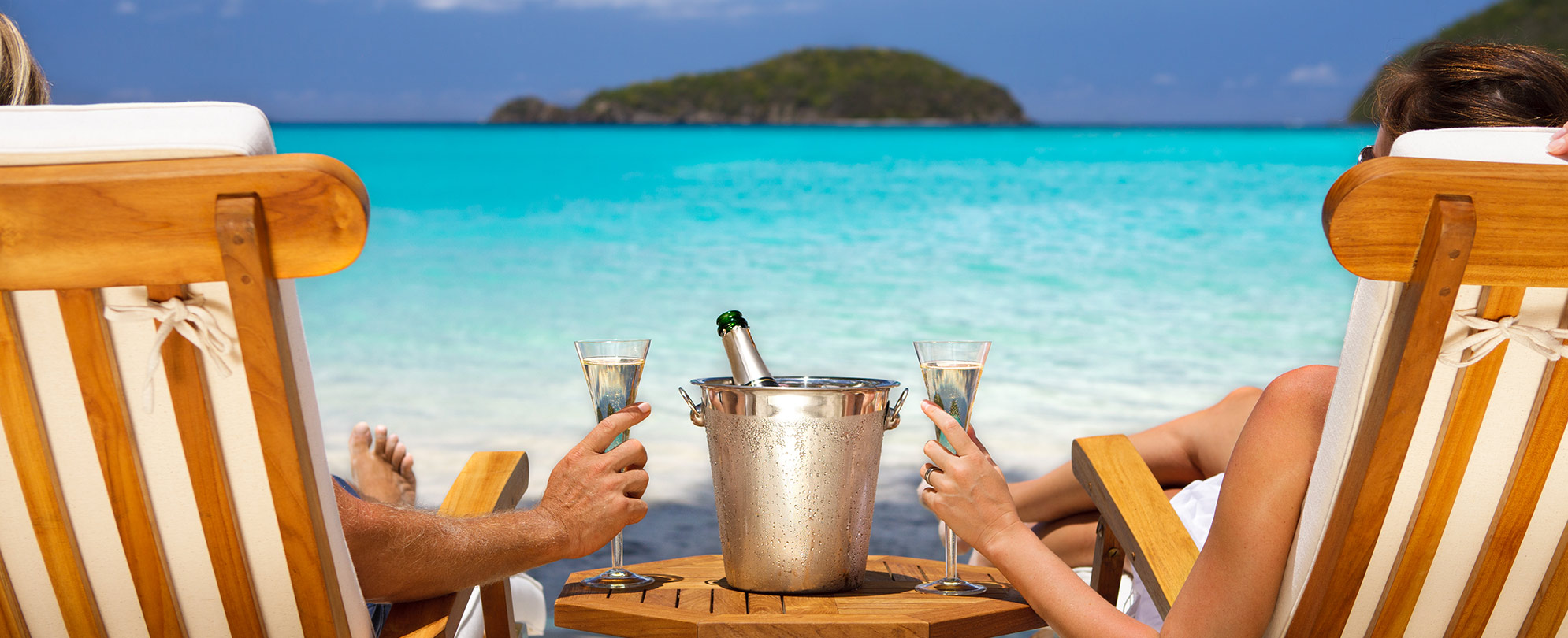 View of a man and a woman from behind as they lounge in beach chairs with a bottle on ice between them and each holding a glass of champagne as they look out at the ocean.