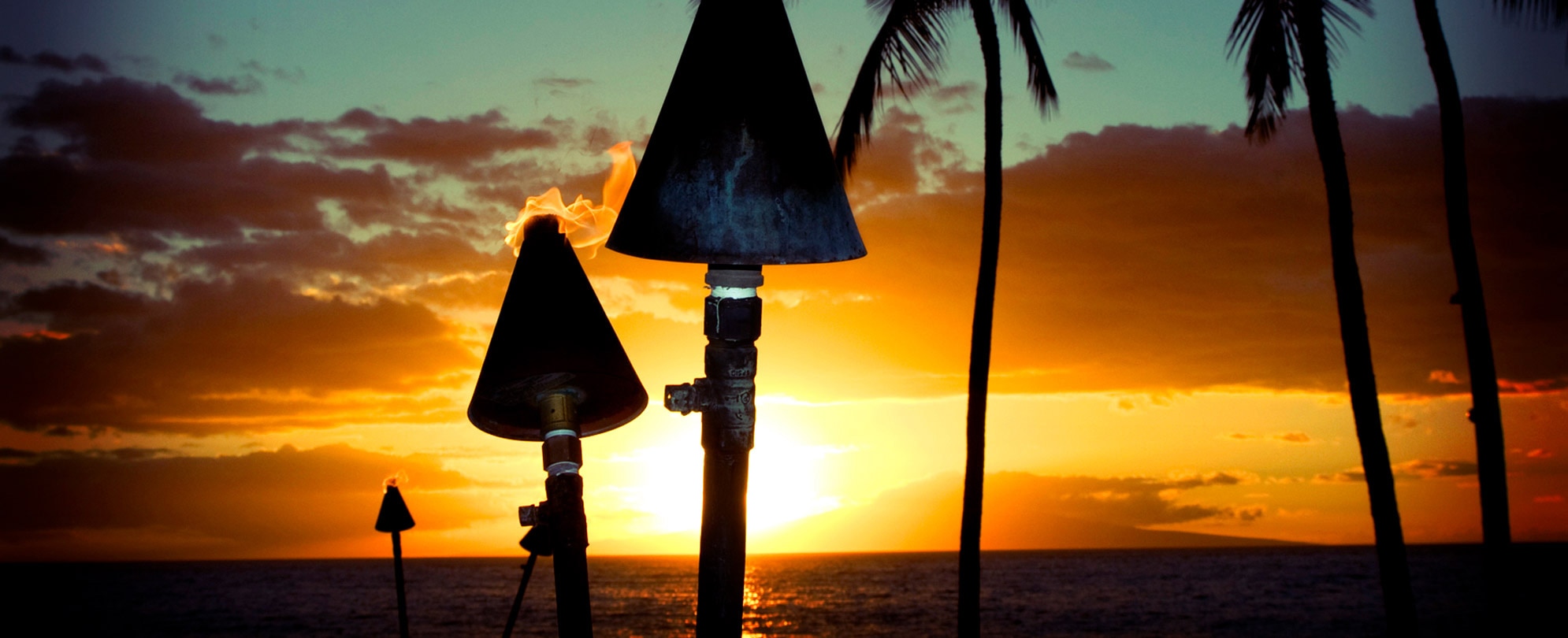 Three tiki torches light up the spooky beach and palm trees at sunset in Hawaii