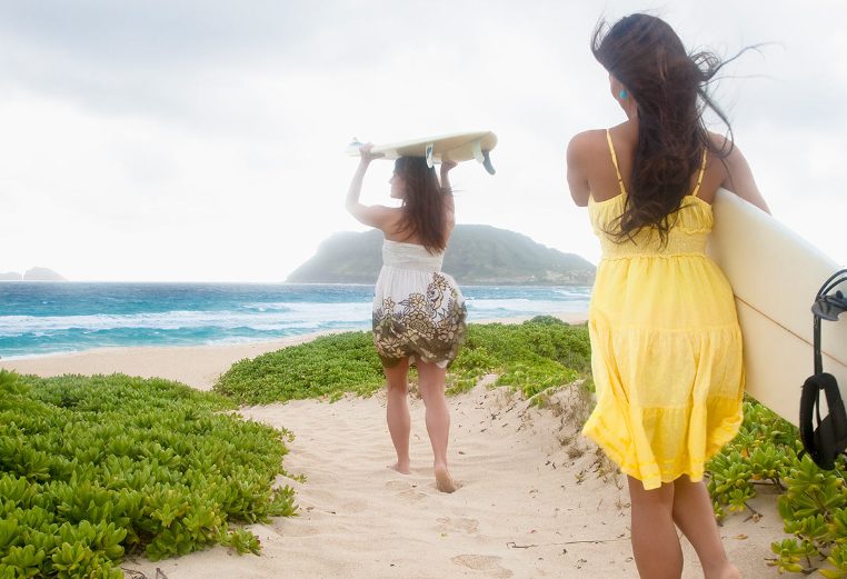 Two women surfers wearing sundresses carry surfboards down a sandy path towards the ocean in Hawaii.